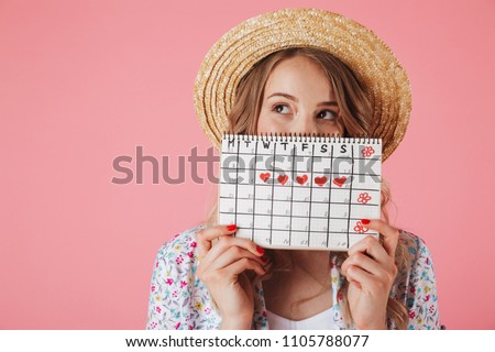 Portrait of a pretty young woman in straw hat holding women's period calendar and looking away isolated over pink background Royalty-Free Stock Photo #1105788077