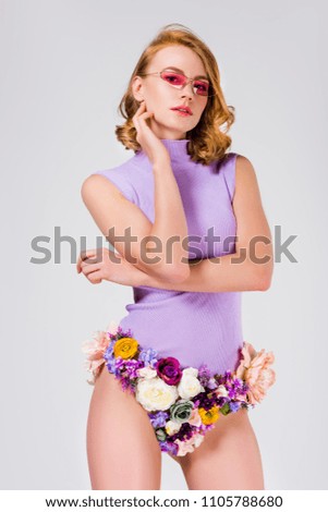 beautiful young woman in sunglasses and panties made of flowers looking at camera isolated on grey