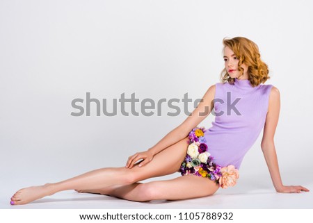 beautiful barefoot girl in panties made of flowers sitting and looking away on grey