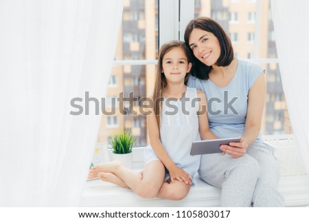 Portrait of beautiful female with dark hair, dressed in casual clothes, holds digital tablet, sits near her daughter, watch something, sit on window sill, play in bedroom. People, lifestyle and family