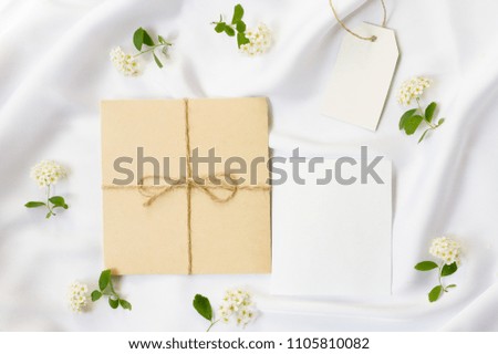 Flat lay workspace, mockup. Wedding invitation cards, craft envelopes,white flowers, green leaves and lace on white satin background. Top view.