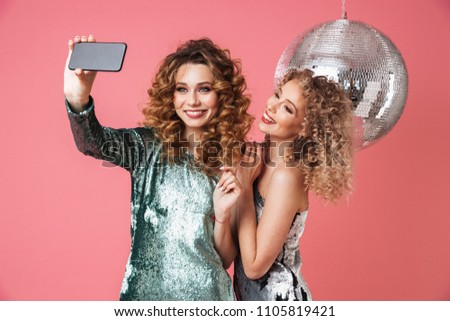 Two beautiful smiling women in shiny dresses taking a selfie while standing with disco ball isolated over pink background
