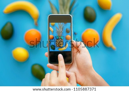 hands holding phone and taking photo of pineapple in sunglasses and bananas, oranges and avocado on blue paper, trendy flat lay. stylish food photography. instagram fruit photo