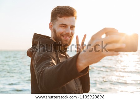 Happy young sportsman taking selfie with mobile phone while standing at the beach and showing peace gesture
