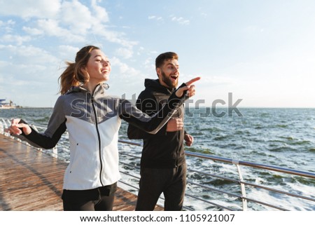 Cheerful young sports couple jogging together at the beach and pointing away