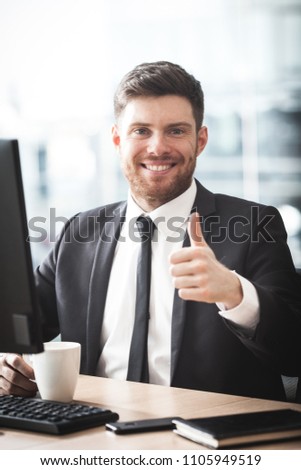 Young businessman at the office sitting at his desk and showing thumb-up sign