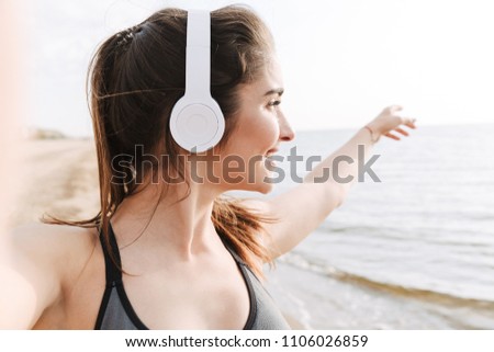 Smiling young sportswoman with headphones taking a selfie with outstretched hands while standing at the beach and pointing away