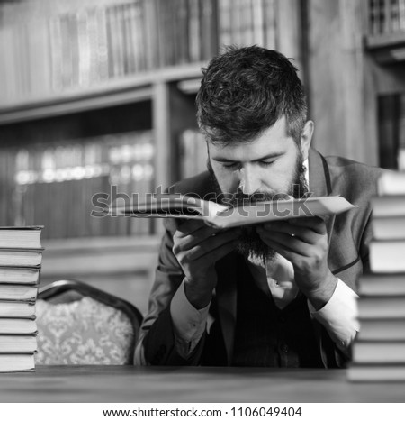 Man with beard in classic suit, scientist or professor sniffs smell of antique book in library near pile of books, bookshelves on background, defocused. Treasures of science concept.