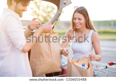 Happy young couple loading grocery bags into a car trunk at a parking lot in front of a shopping mall. Royalty-Free Stock Photo #1106055488