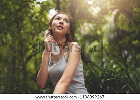 Asian Beautiful woman in grey vest looking up and holding a bottle of water on her neck while sitting over green forest background. Thirsty, Resting. Smiling Face. Freshness.