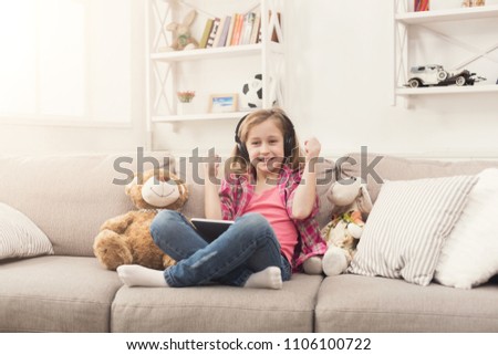 Little girl playing online games on digital tablet and listening to music in headphones. Female child sitting on sofa with her toys. Social networking and online education concept