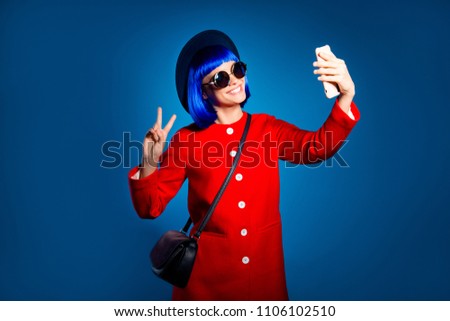Portrait of positive toothy girl having video-call shooting self portrait on front camera of smart phone gesturing hi v-sign with hand, isolated on bright blue background with light and shadow