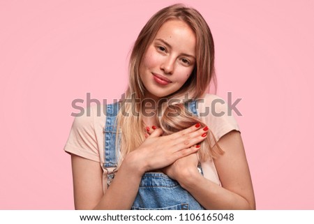 Pleased lovely Caucasian female with friendly look, keeps hands on heart, demonstrates her good attitude, dressed in casual t shirt and denim overalls, expresses sincere feelings, has red manicure
