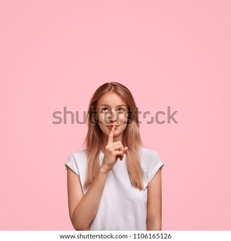 Pretty European young female shows silence sign and asks to keep private information confidential, looks thoughtfully and secretly upwards, wears oversized white t shirt, isolated over pink background Royalty-Free Stock Photo #1106165126