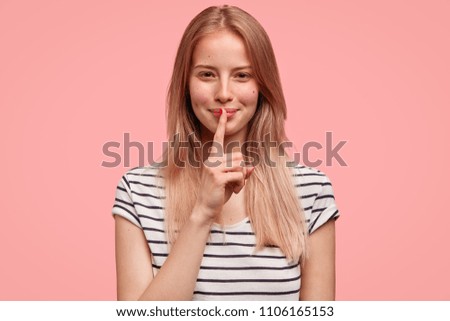 Caucasian female model with positive expression, keeps fore finger on lips, wears striped t shirt, isolated over pink wall. Secret woman gossips with friend, tells private information, makes hush sign