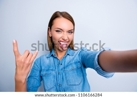 Self portrait of cool crazy girl shooting selfie on front camera showing rock and roll symbol with tongue out isolated on grey background