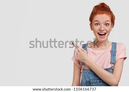 Freckled young foxy female in casual outfit, has amazed look, indicates with fore finger at right upper corner, shows copy space for your information, look with suprisement and happiness at camera Royalty-Free Stock Photo #1106166737
