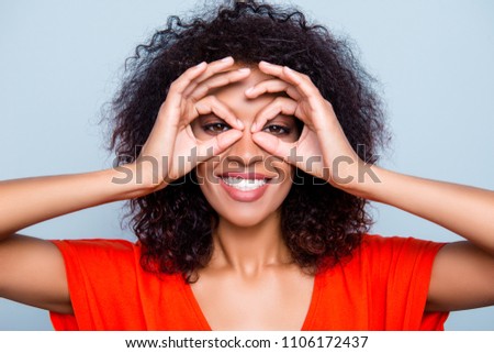 Closeup portrait of joyful toothy woman with modern hairdo in orange outfit making binoculars with fingers looking at camera isolated on grey background