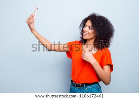 Portrait of cheerful trendy model in vivid outfit with modern hairdo shooting selfie on front camera of smart phone gesturing thumb up like sign with finger isolated on grey background
