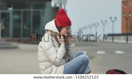 Young beautiful woman in a red hat wearing sporty warm clothes and rollers, sitting on the asphalt road and talking on the phone 4k