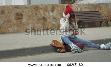 Young beautiful woman in red hat wearing sporty warm clothes and rollers, sitting on the asphalt road and taking pictures on a vintage camera 4k