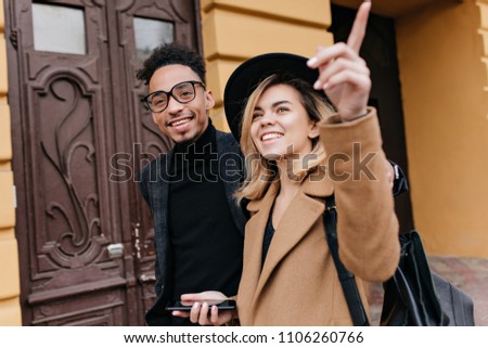 Carefree blonde woman in hat showing her african friend something interesting. Outdoor portrait of smiling black guy in glasses walking around town in cold day with fair-haired lady.