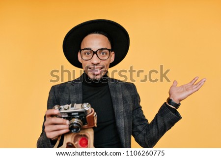 Studio shot of surprised male photographer in black hat. Indoor photo of african young man with camera isolated on yellow background.