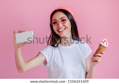 Young asian teenage girl isolated on pink background. Stylish young woman with ice cream is making selfie on a smart phone.
