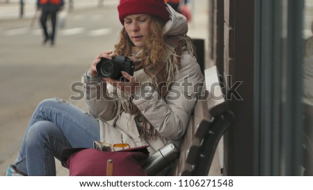 Young beautiful woman in a red hat wearing sporty warm clothes and rollers, sitting on a wooden bench and taking pictures on a vintage camera 4k
