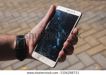 A man's hand holds a mobile phone with a broken screen on the background of the paving stones on a clear day. Broken smartphone. Small shards. Black smashed screen. Unlucky day. Insurance. Watch strap