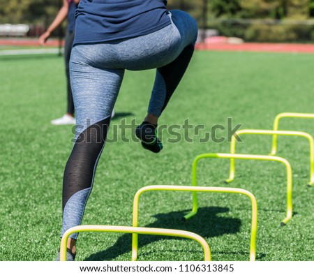 A female high school athlete performs running drills over yellow mini banana hurdles on a turf field with no shoes on, only socks in blue spandex. Royalty-Free Stock Photo #1106313845