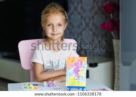 a little preschool girl sits in a room with a table with paints and a picture