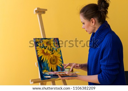 Girl artist paints sunflowers oil paints on canvas. She is wearing blue sweater. Woman is holding brush and palette with paints. She mixes colors with her fingers.