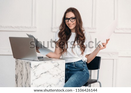 A young student studies in a bright room, just printed out her report on paper and checks it. A happy girl in fashionable glasses smiles and holds sheets of paper in her hands.