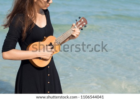 Woman wearing black dress with ukulele standing on the beach of sea background.