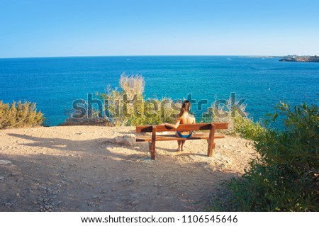 Image of a back of one tan caucassian girl with wet hair sitting on wooden bench with her hand leaning behind, looking in the distance to the blue sea. Cloudless summer day