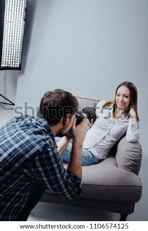 Professional photo shoot at the studio, a female model is posing on an armchair and smiling; the photographer is taking pictures with his digital camera