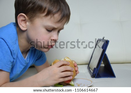cute boy at home on the couch, eating a hamburger and looking at the tablet, watching cartoons or talking with friends he eat burgers and look at something on the tablet. he focused on screen