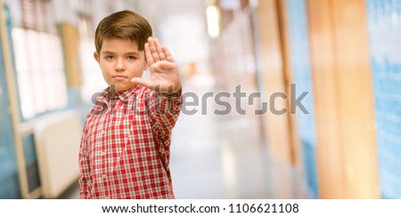 Handsome toddler child with green eyes annoyed with bad attitude making stop sign with hand, saying no, expressing security, defense or restriction, maybe pushing at school corridor