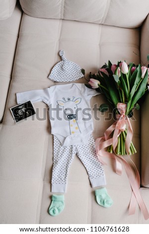 Set of baby clothes in white color. Newborn accessories: socks, cap, ultrasound scan or picture of boy. Bouquet flowers greetings for a party in honor of pregnant at home. Top rear view. baby shower.