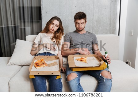 Unhappy couple ate too much pizza while sitting together on a couch at home Royalty-Free Stock Photo #1106786201