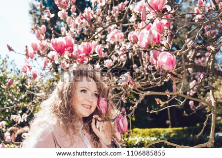 curly happy young woman posing next to a blooming pink magnolia, laughing and enjoying the warmth