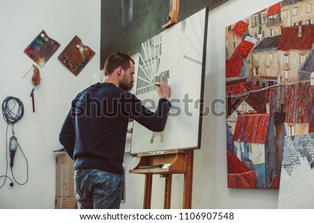 Concentrated male artist is drawing a dog on easel in his art studio, lots of pictures, creative atmosphere