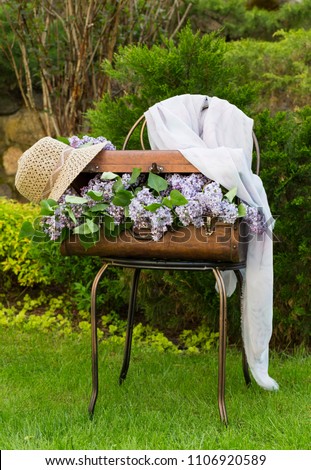 on the old chair is a suitcase, in it a bouquet of purple lilac, a straw hat and a shawl. The composition is located in the garden on the grass