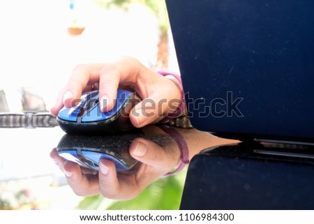 Female hand on computer mouse. The blue mouse. On the glass table. Blurred background. A laptop. An object. Subject. Close-up, macro. Street, greenery.