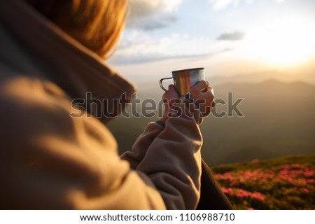 Closeup photo of cup with tea in traveler's hand over out of focus mountains view. A young tourist woman drinks a hot drink from a cup and enjoys the scenery in the mountains. Trekking concept