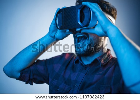 Casual bearded man wearing VR goggles and looking shocked and amazed in blue neon light