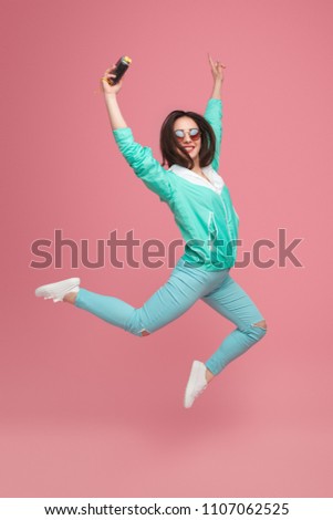 Trendy brunette in blue stylish outfit and sunglasses jumping on pink background holding retro camera in hand