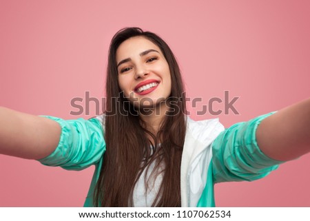 Pretty young girl with red lipstick and in stylish sportive jacket smiling at camera taking selfie on pink background