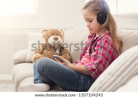 Happy little casual girl in headphones listening to music on smartphone. Pretty kid at home watching cartoon on mobile phone online, sitting on sofa with her teddy bear, copy space